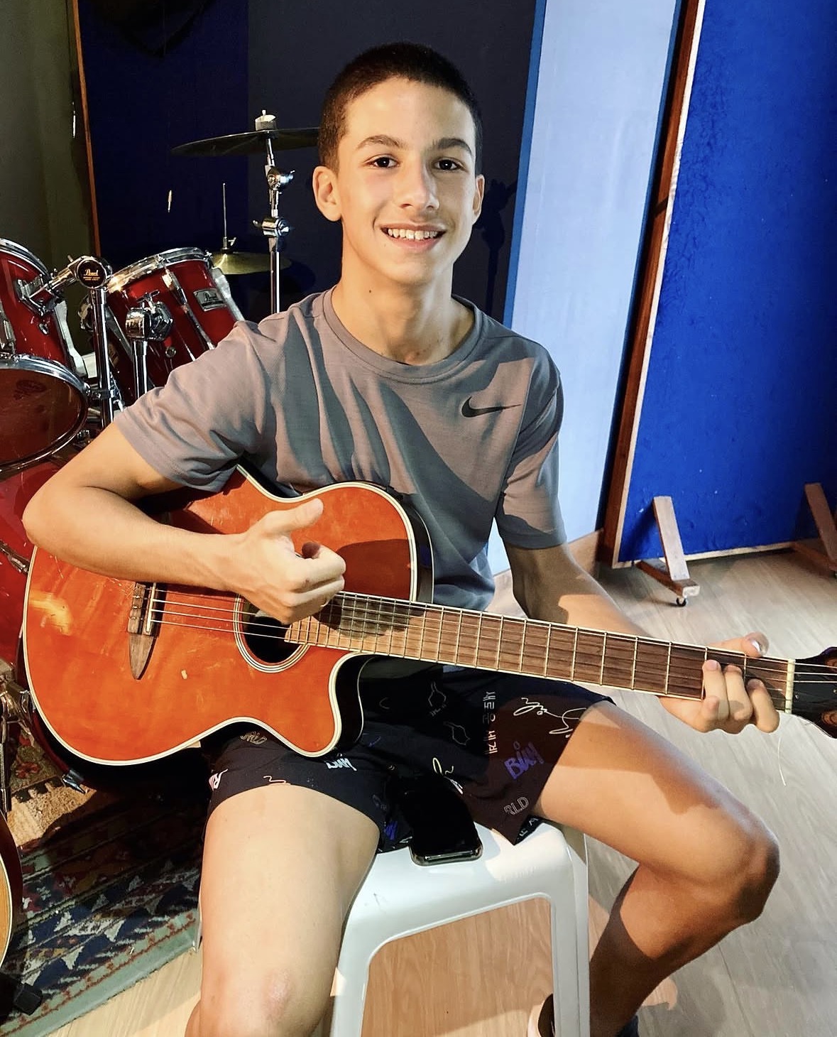 Adolescente sorrindo com violão em estúdio, com bateria vermelha ao fundo