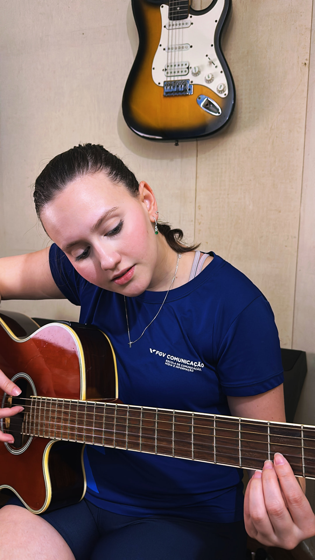 Aluna tocando violão acústico na sala com guitarras na parede